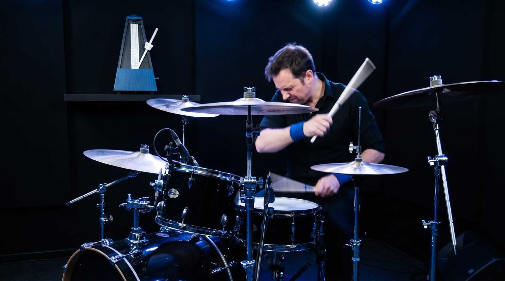 Drummer performing on an acoustic drum kit beside a metronome in a dark studio.