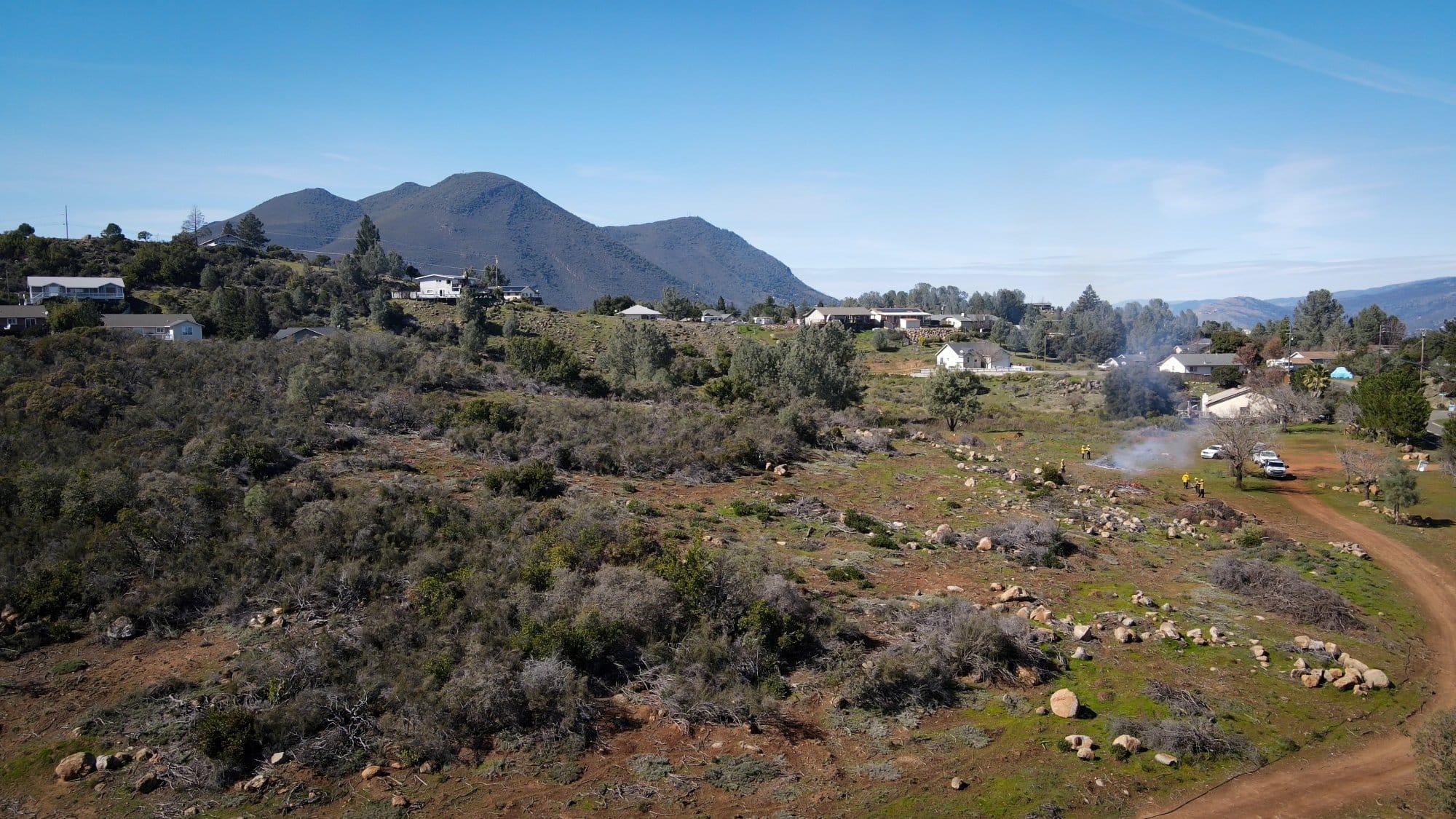 Homes on a hillside at the edge of Kelseyville, California, sit next to a wide field of brush and chaparral, where a fuels crew and a thin line of smoke are visible in the distance.