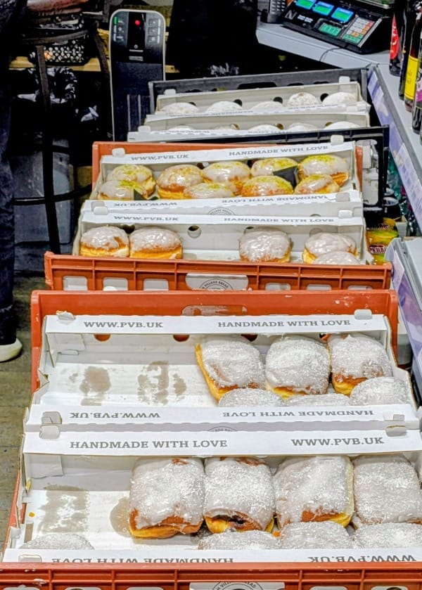 A line of  rust-red coloured plastic bakers trays behind the till counter filled with different kinds of doughnuts. The ones in the foreground are the folded cheese doughnuts. The rest are either dusted with sugar or covered in white icing. Some of the iced doughnuts are sprinkled with pistachios