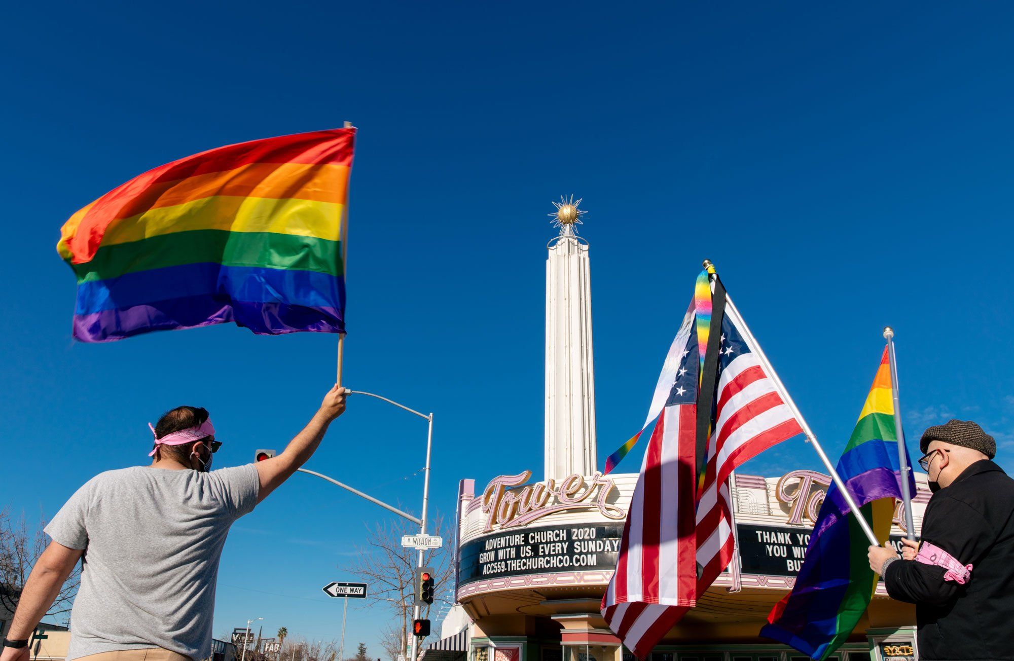 An individual waves a rainbow flag next to another individual holding an American and rainbow flag, across the street from the Tower District Theatre. The Tower Theatre can be seen in the background displaying a marquee advertising Adventure Church’s message, “Adventure Church 2020. Grow with us, every Sunday” along another marquee displaying, “Thank you Rush for teaching us, You will be missed very much but never forgotten. RIP.”