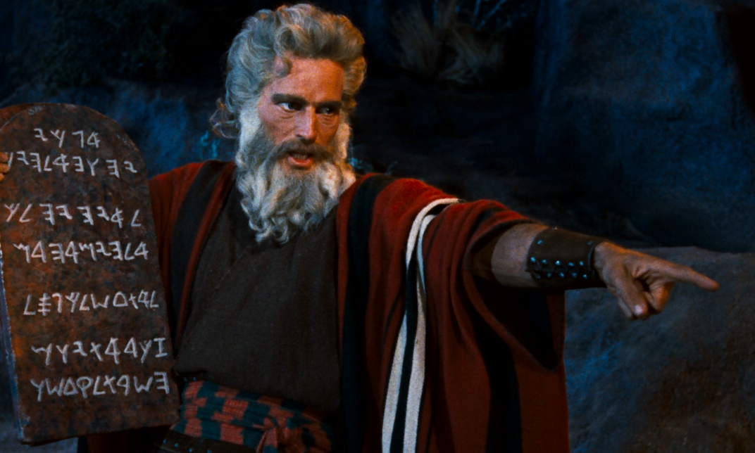 A still from the film "The Ten Commandments" starring Charleton Heston as Moses. The image is of Moses holding one tablet with writing on it