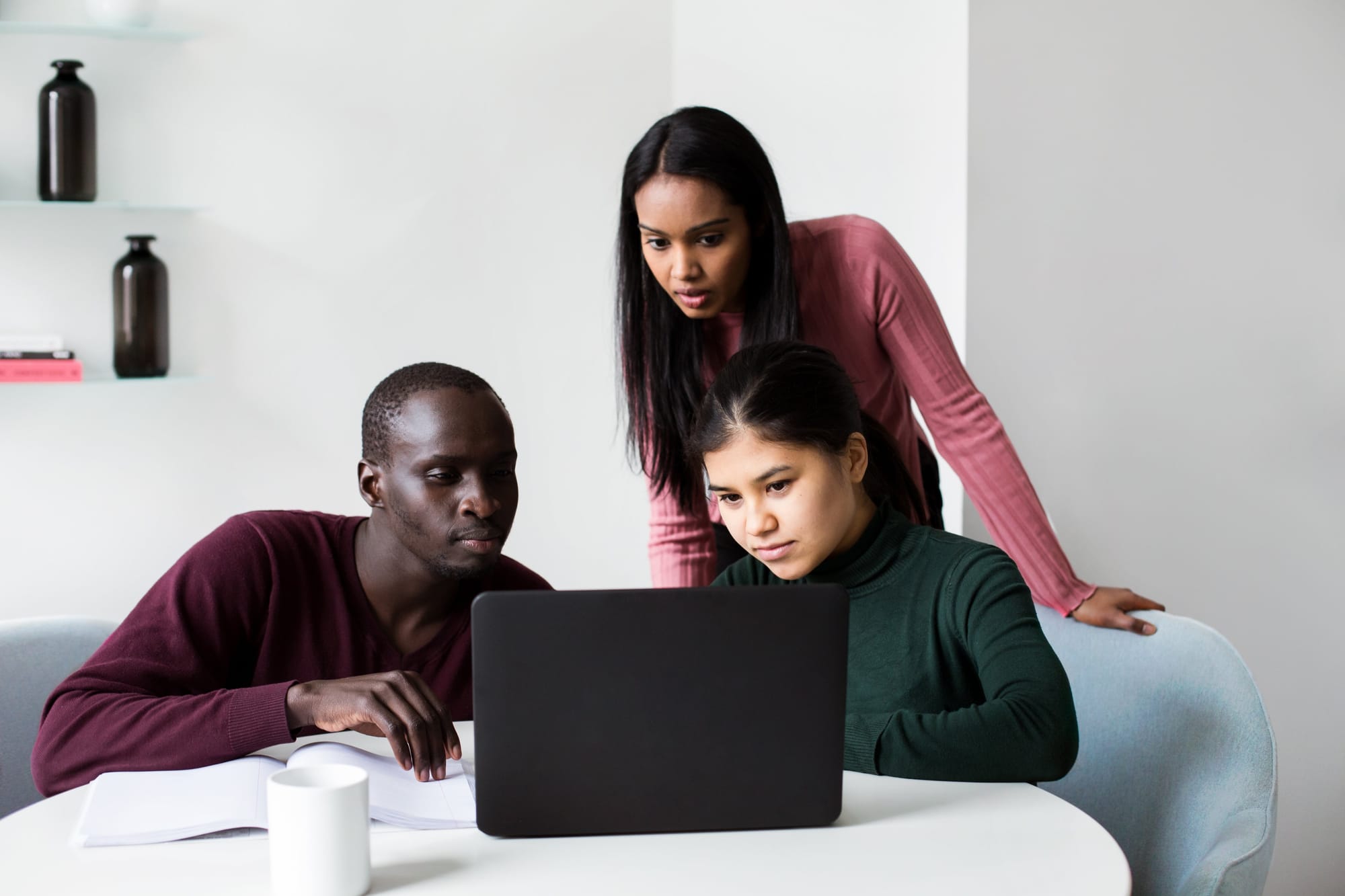 Three colleagues gathered around a laptop at a table, two seated and one leaning over to look at the screen.