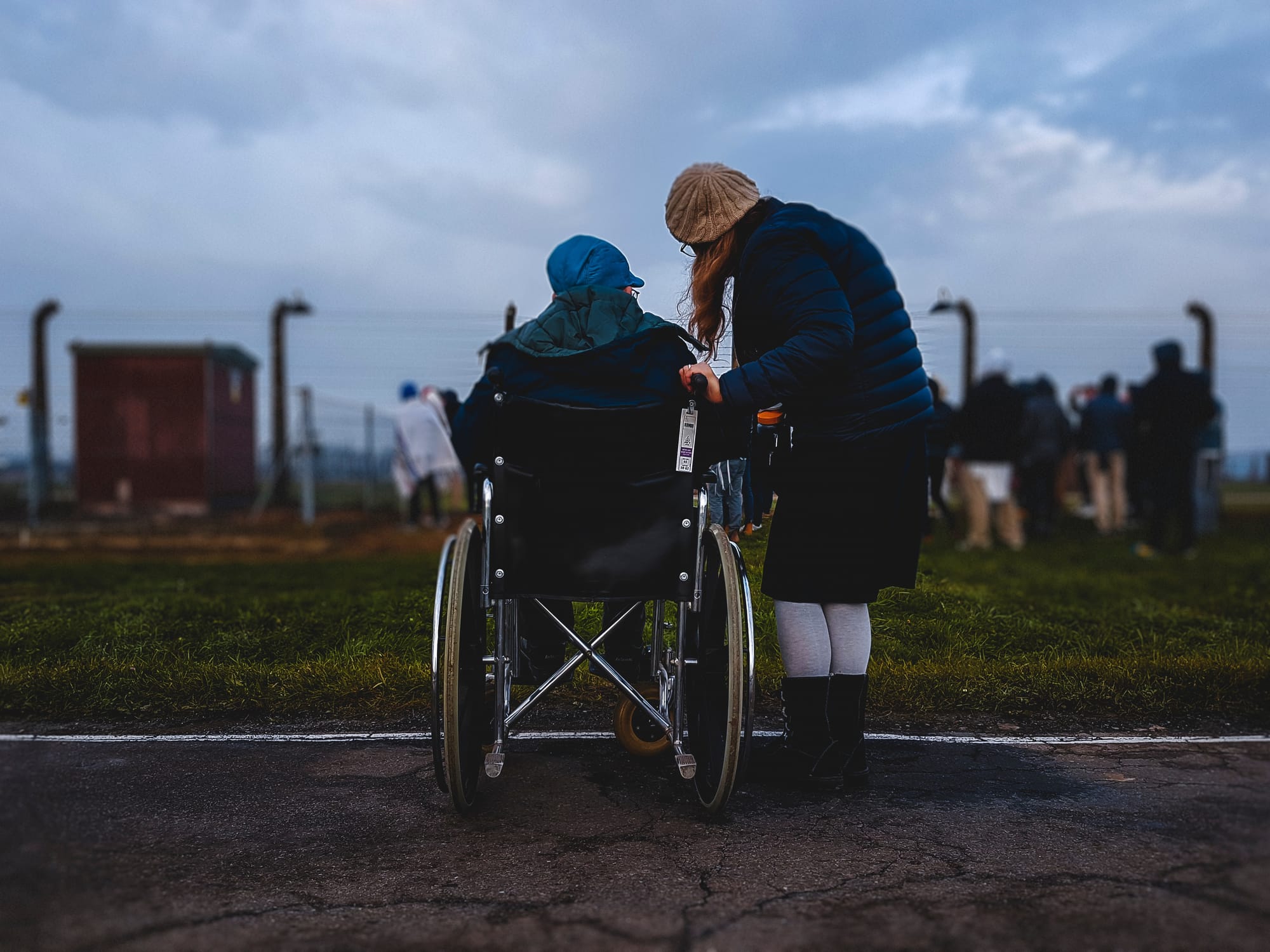Seen from behind, one person leans in to talk with another seated in a wheelchair on an open road under an overcast sky.