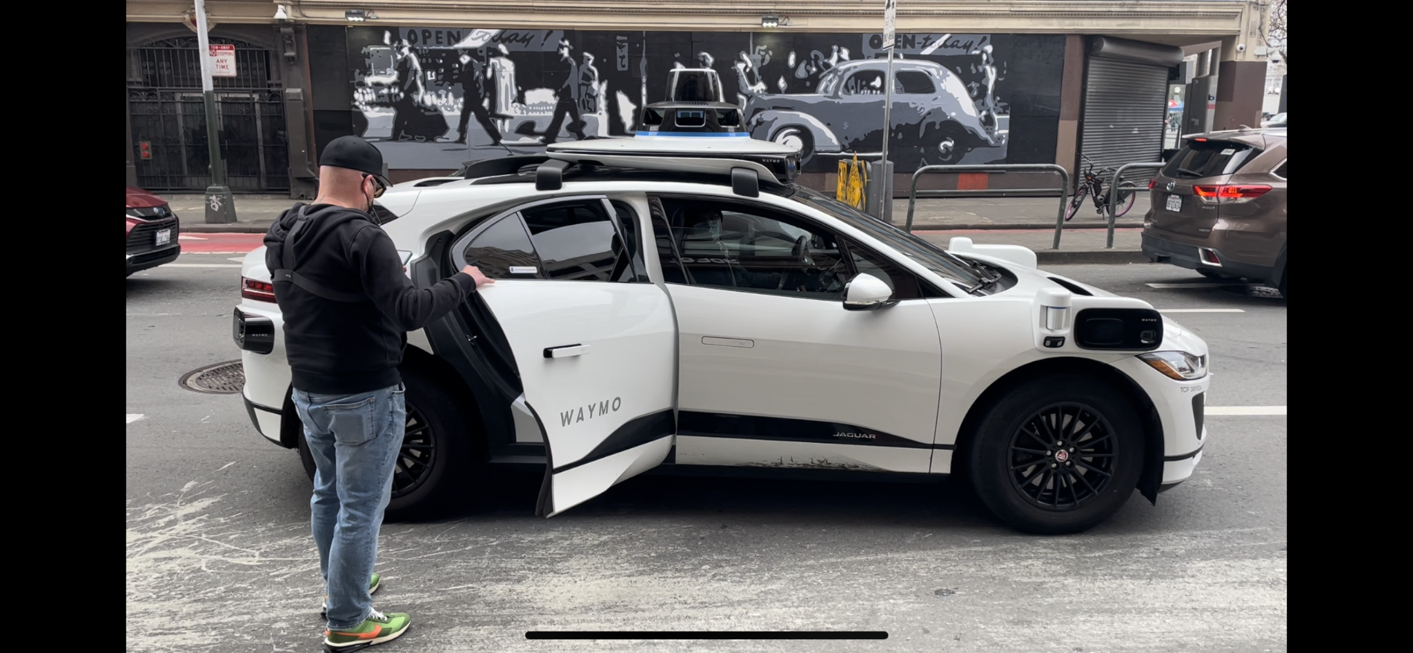 A participant opening the rear door of a white Waymo vehicle at the curb.