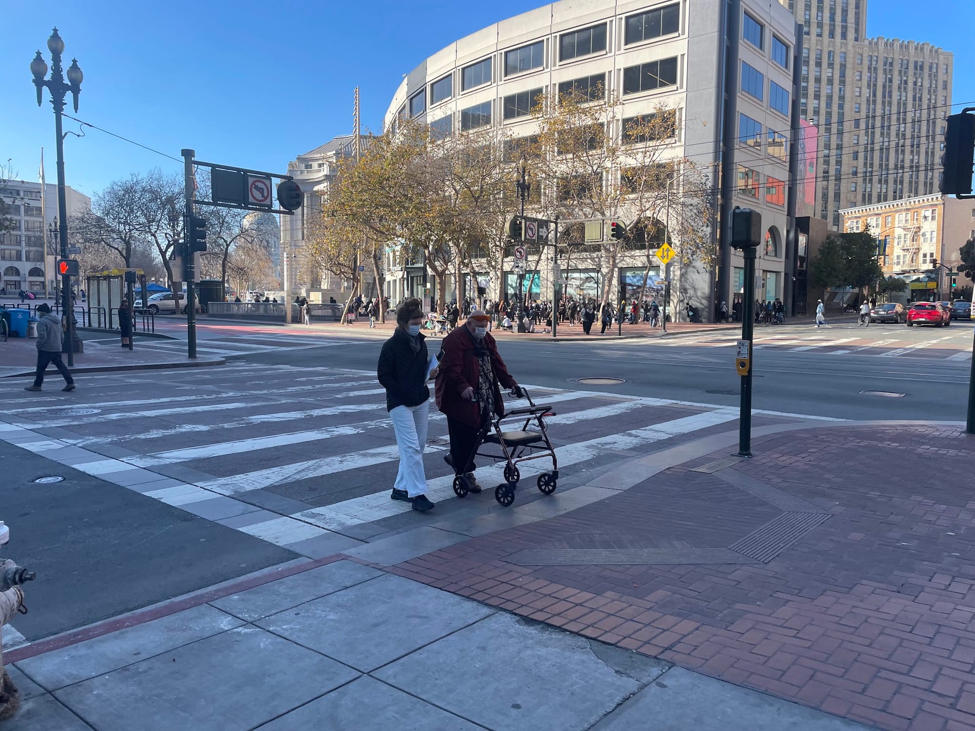 Two people crossing a sunny downtown San Francisco intersection, one using a walker.
