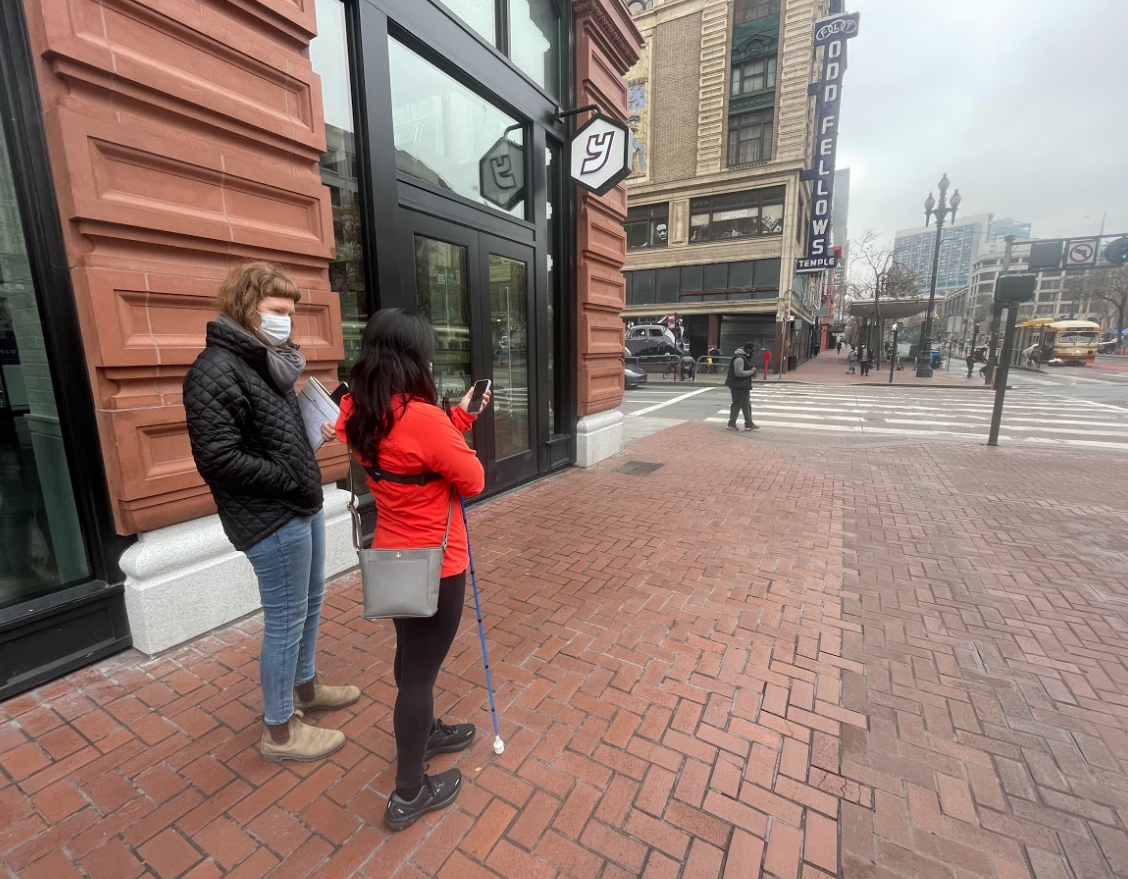 A participant in a red jacket with a white cane checks a phone beside a researcher on a brick sidewalk.
