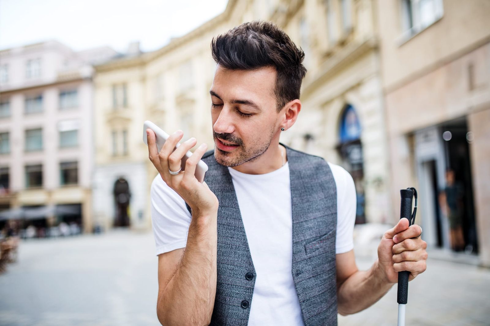 A man with a white cane holds a smartphone to his ear listening to a screenreader while standing in a city square, wearing a white t-shirt and a grey button-up vest over it.