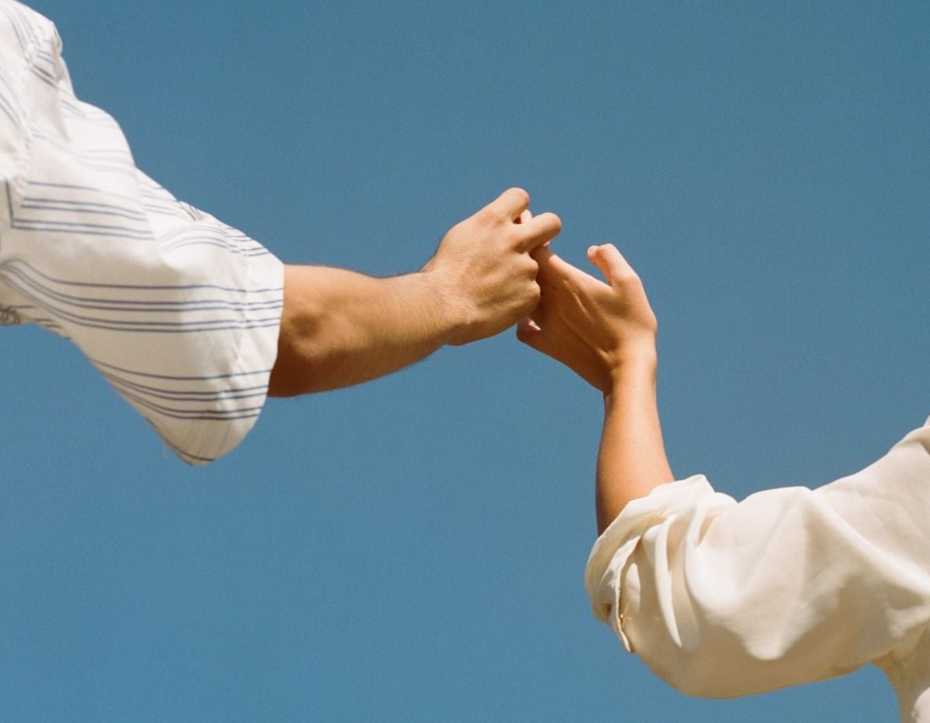 A clear blue sky frames two people in white blouses holding hands, only their shoulders and arms in shot, focusing on the connection between them.