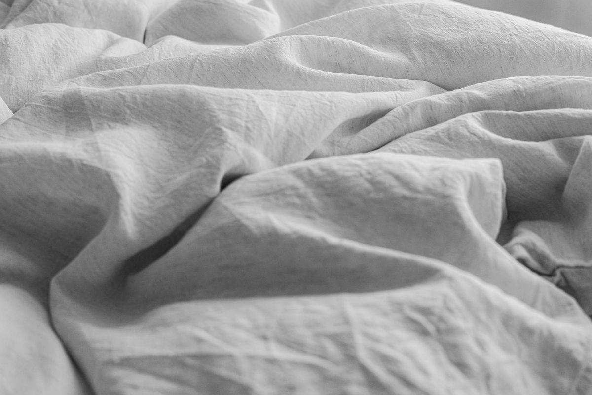 A close-up photograph of rumpled, textured white linen sheets on a bed, captured in soft, natural morning sunlight and shadows.