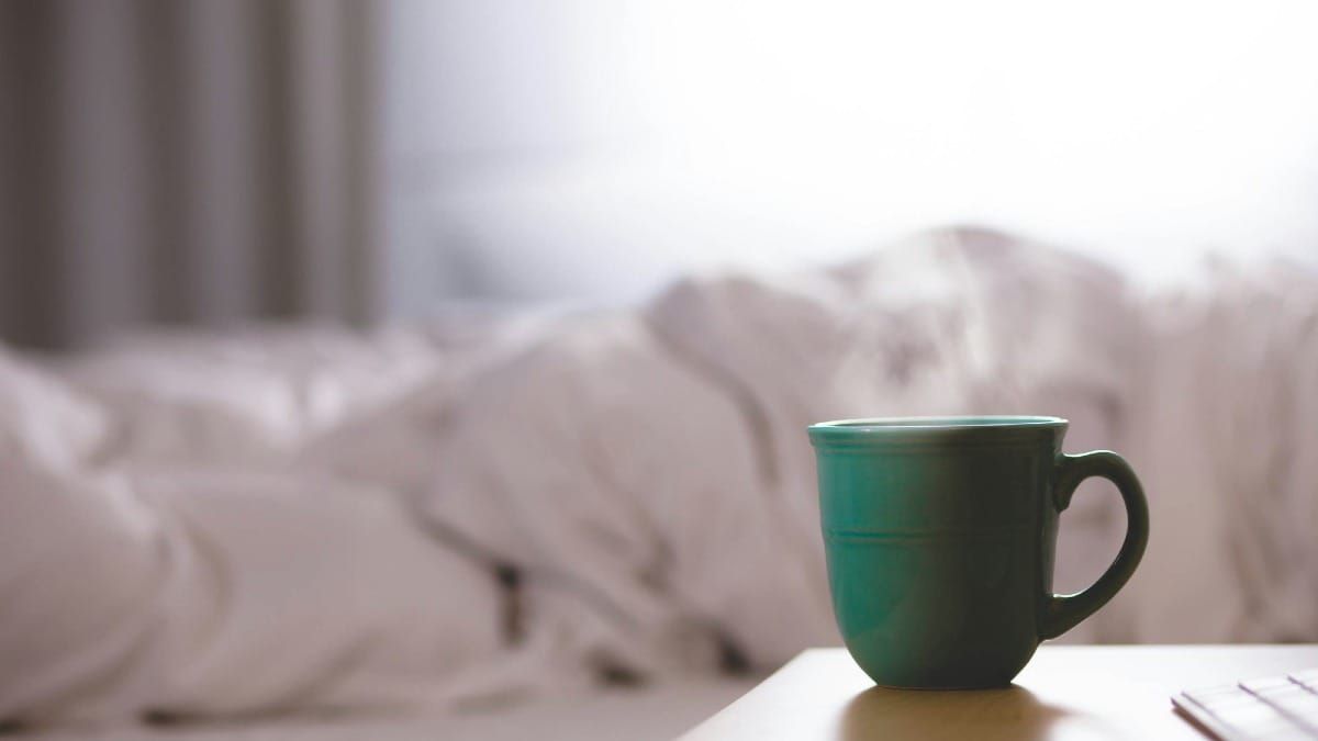 A close-up photograph of a steaming green ceramic mug sitting on a wooden table next to a bed with rumpled white linens.