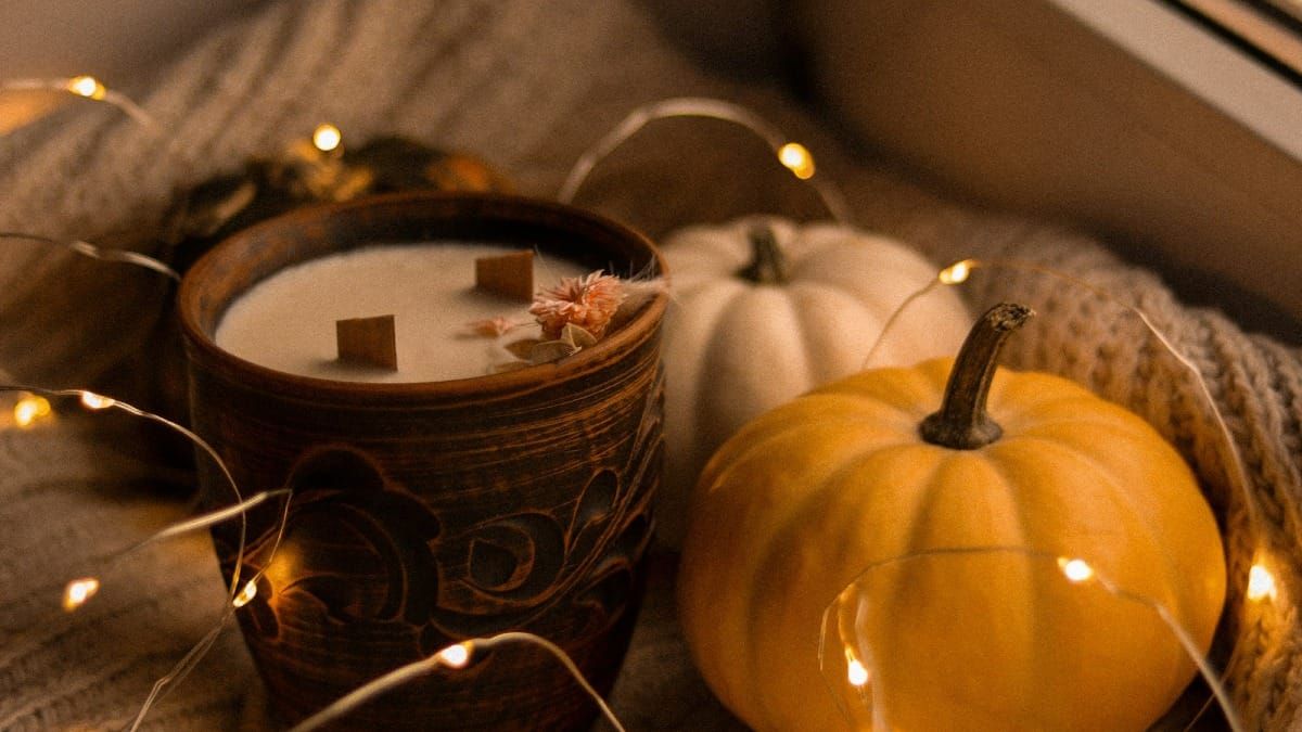 A cozy, close-up photograph of small yellow and white autumn pumpkins sitting next to a rustic ceramic wood-wick candle wrapped in warm, glowing fairy lights.