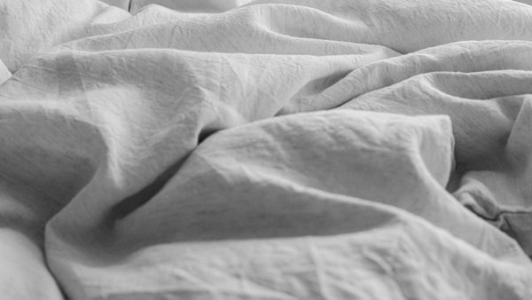 A close-up photograph of rumpled, textured white linen sheets on a bed, captured in soft, natural morning sunlight and shadows.