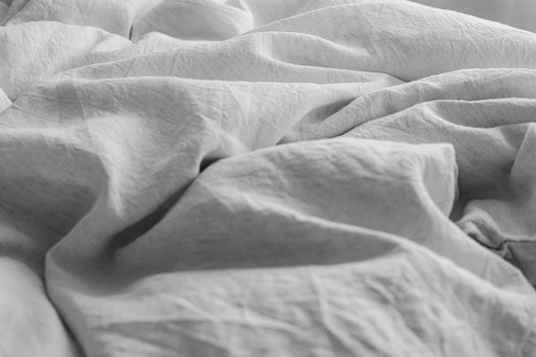 A close-up photograph of rumpled, textured white linen sheets on a bed, captured in soft, natural morning sunlight and shadows.