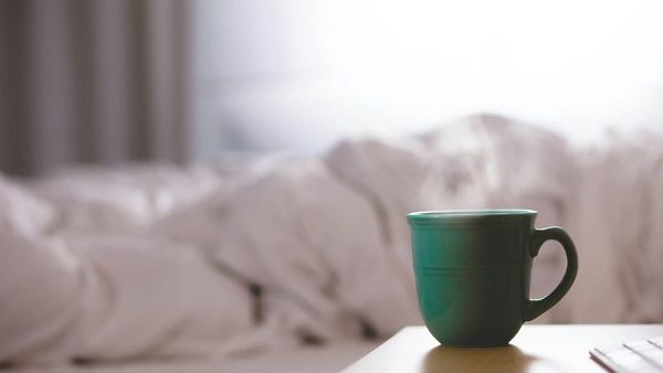 A close-up photograph of a steaming green ceramic mug sitting on a wooden table next to a bed with rumpled white linens.