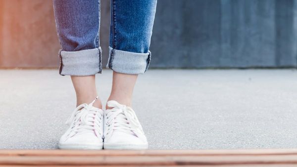 A close-up photograph of a person wearing cuffed blue jeans and white sneakers standing on a concrete surface next to a wooden bench.