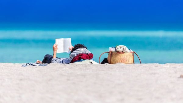 A photograph from behind of a person lying on a sandy beach reading an open book, next to a woven tote bag with a vibrant blue ocean in the background.