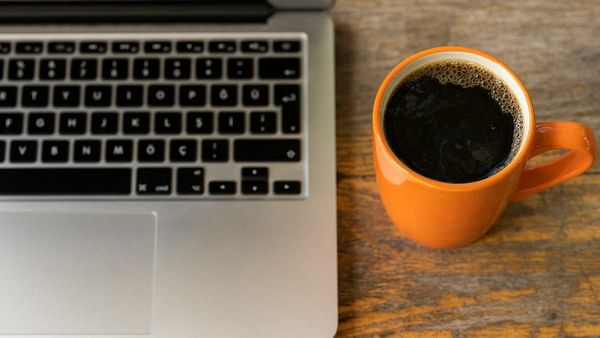 n overhead photograph of an open silver laptop sitting on a rustic wooden table next to an orange ceramic mug filled with black coffee.