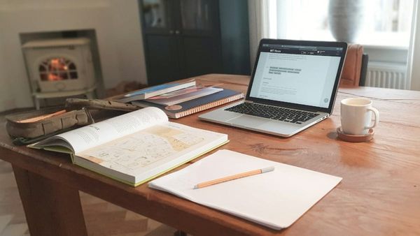 A cozy indoor workspace on a large wooden table featuring an open laptop, a white coffee mug, notebooks, an open book, and a warm fireplace burning in the background.