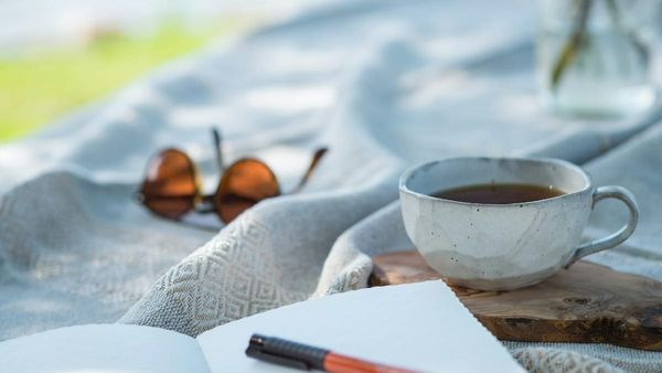 A close-up photograph of an open notebook and pen on a textured linen blanket next to a ceramic coffee mug and a pair of sunglasses outdoors.