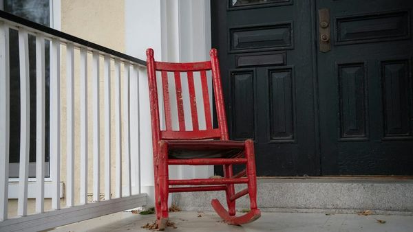 A photograph of a weathered red rocking chair sitting on a house front porch next to a white railing and a closed dark front door.