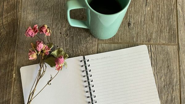 A high-angle photograph of an open lined notebook, a sprig of dried pink roses, and a teal ceramic coffee mug resting on a rustic wooden table.