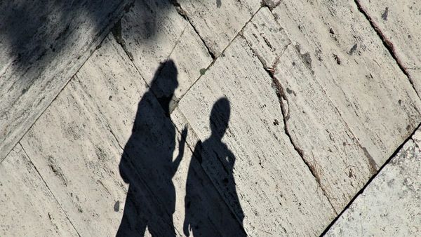 n artistic, high-contrast photograph of the long dark shadows of two people walking side-by-side across textured stone pavement tiles.