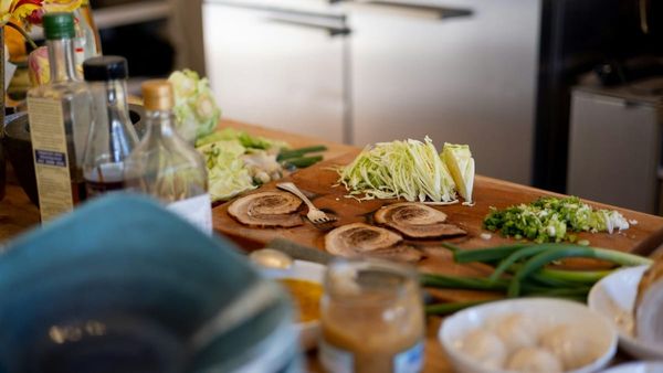 A close-up photograph of a wooden cutting board with sliced green vegetables and pork, surrounded by kitchen bowls and cooking ingredients.