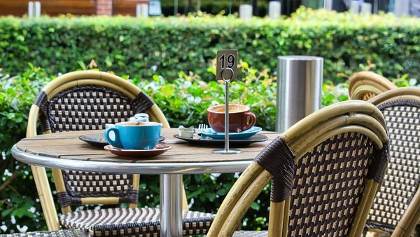 An outdoor café table setting on a sunny day featuring a round wooden table, woven bistro chairs, two coffee cups, and a green hedge background.