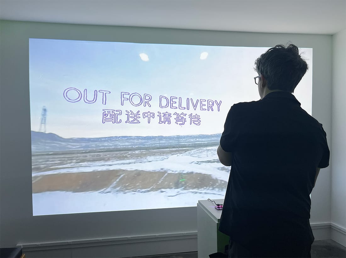A photo of someone playing the documentary game OUT FOR DELIVERY at the SEQUENCEBREAK// exhibition. The game is projected onto a white gallery wall and features a title card featuring a landscape shot of plains. A tower is seen in the distance. The game's title is written in English and Chinese.  