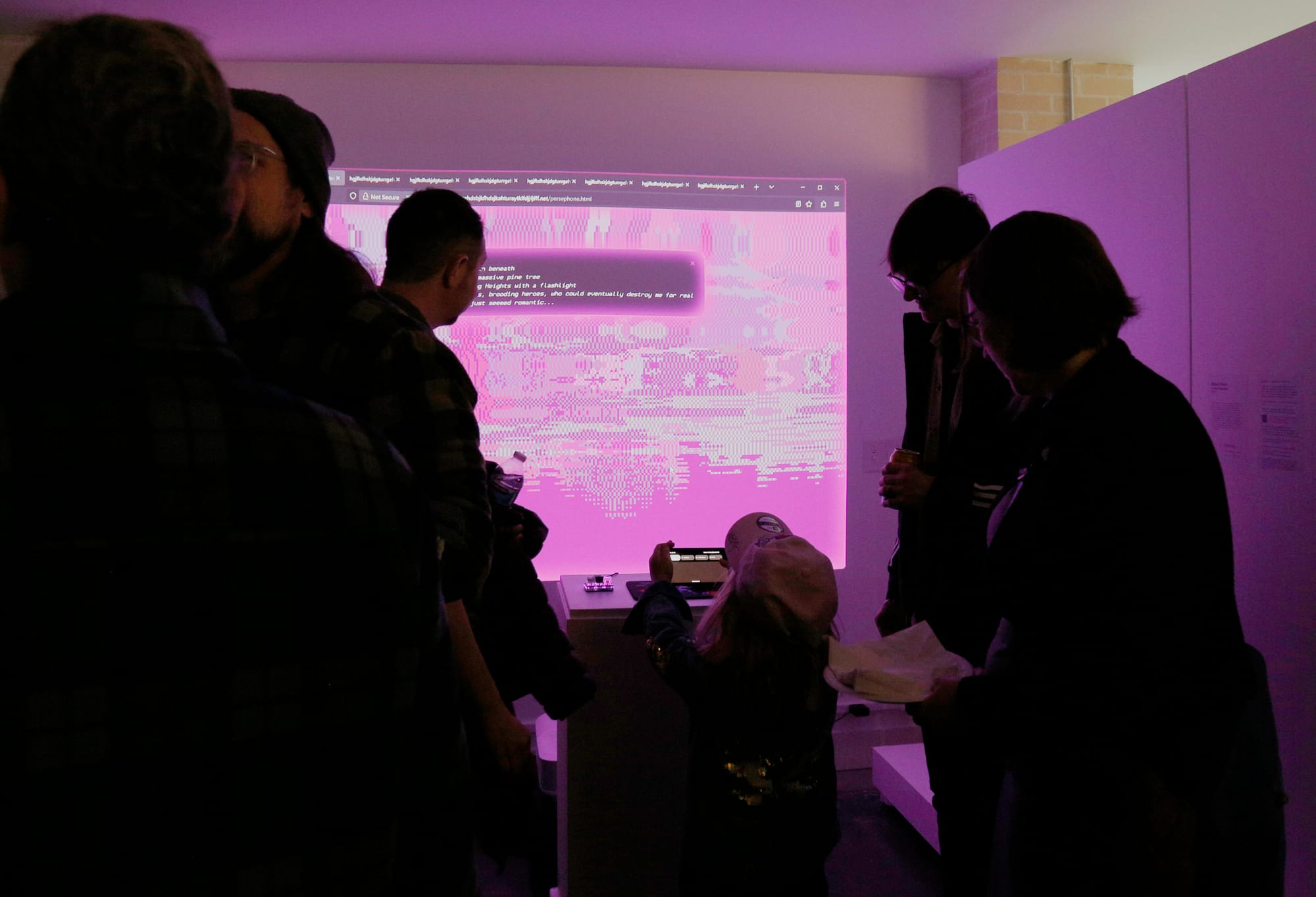 A photograph of Cassie-McQuater's cyberfeminist installation of Black Room, which is projected onto a wall of the gallery space. Several patrons surround the installation, which features retro pixellated graphics and glitch textures. There is a text box on the screen, but the type is too small and blurry to read. 