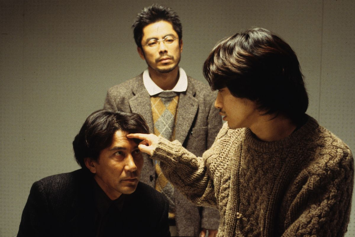 Takabe looks at Mamiya in exasperation as Mamiya pokes Takabe's forehead. Takabe's forensics partner watches Mamiya from behind Takabe.