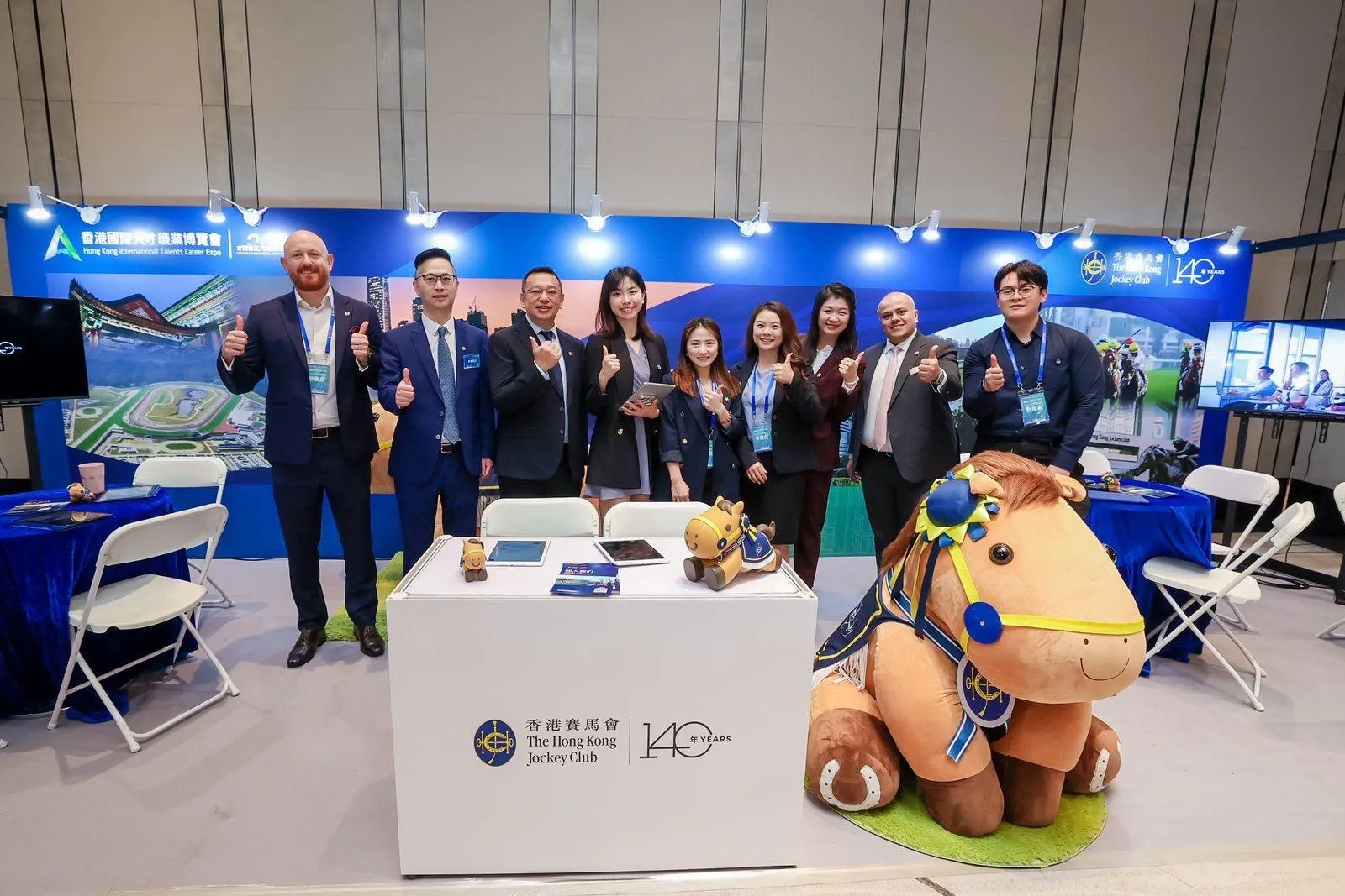 HKJC recruiters in navy suits and blazers line up behind a career-expo booth, all giving thumbs-up beside an oversized plush horse mascot.