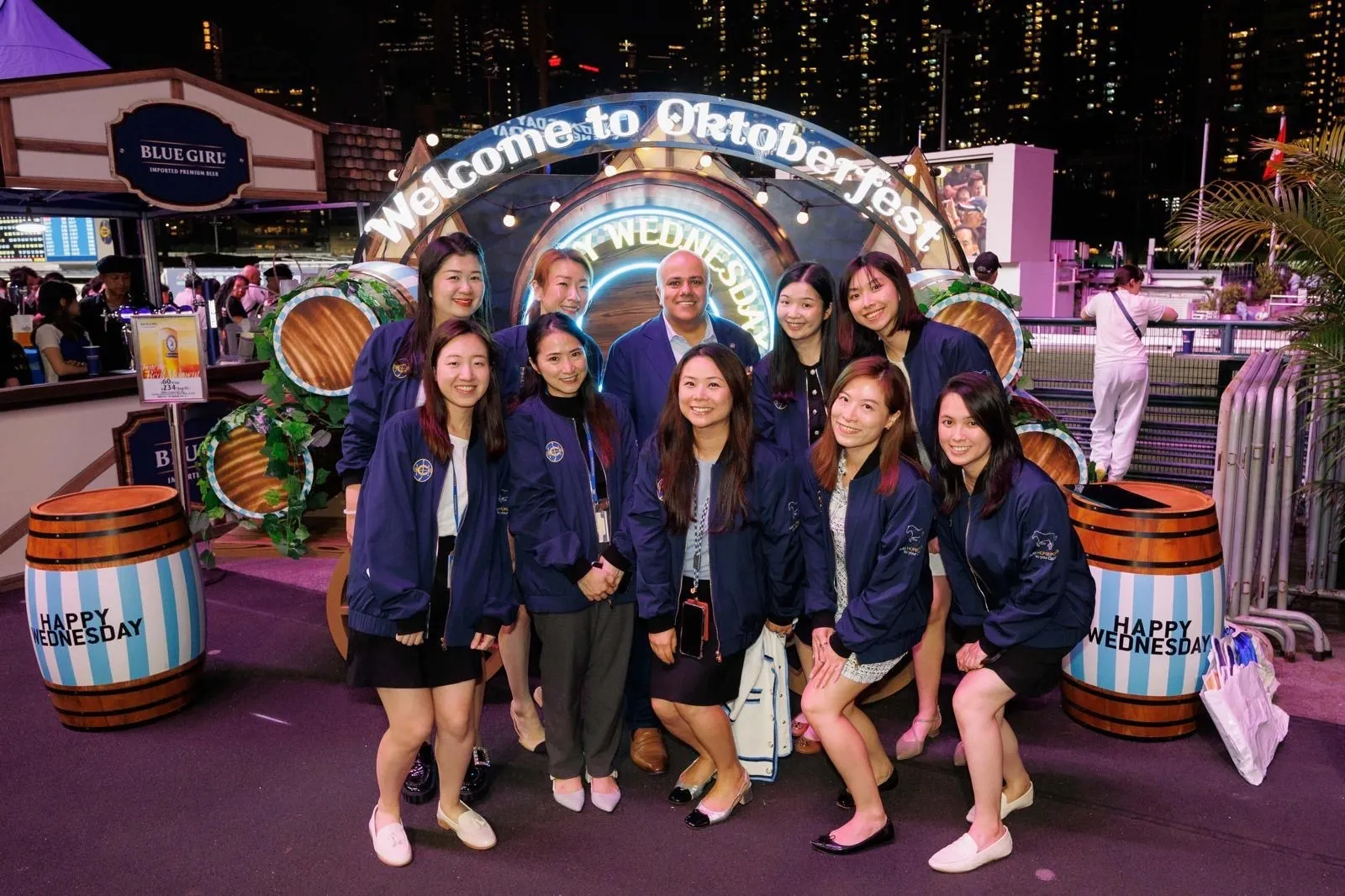 Night-time group shot of HKJC staff in navy jackets beneath a lit “Welcome to Oktoberfest” arch at Happy Wednesday trackside festivities.