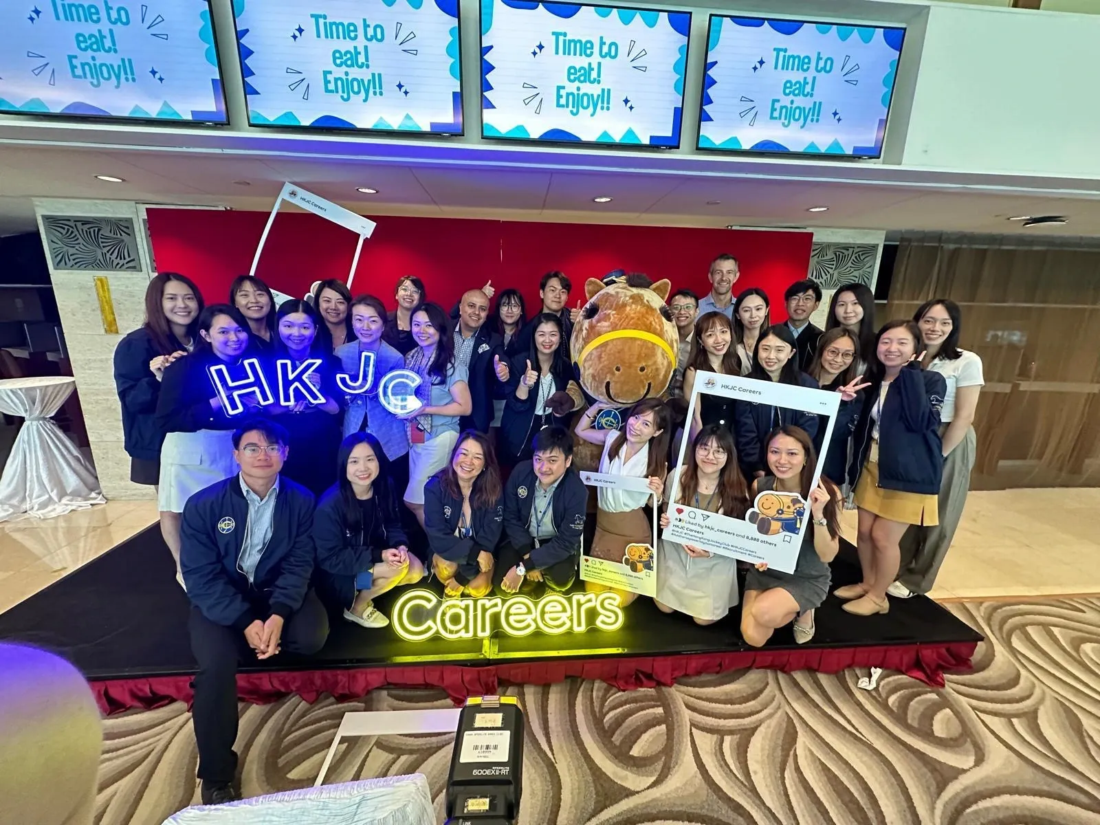 Two dozen young professionals crowd a small stage, posing with a neon “HKJC Careers” sign and a cuddly horse mascot under “Time to eat!” screens.