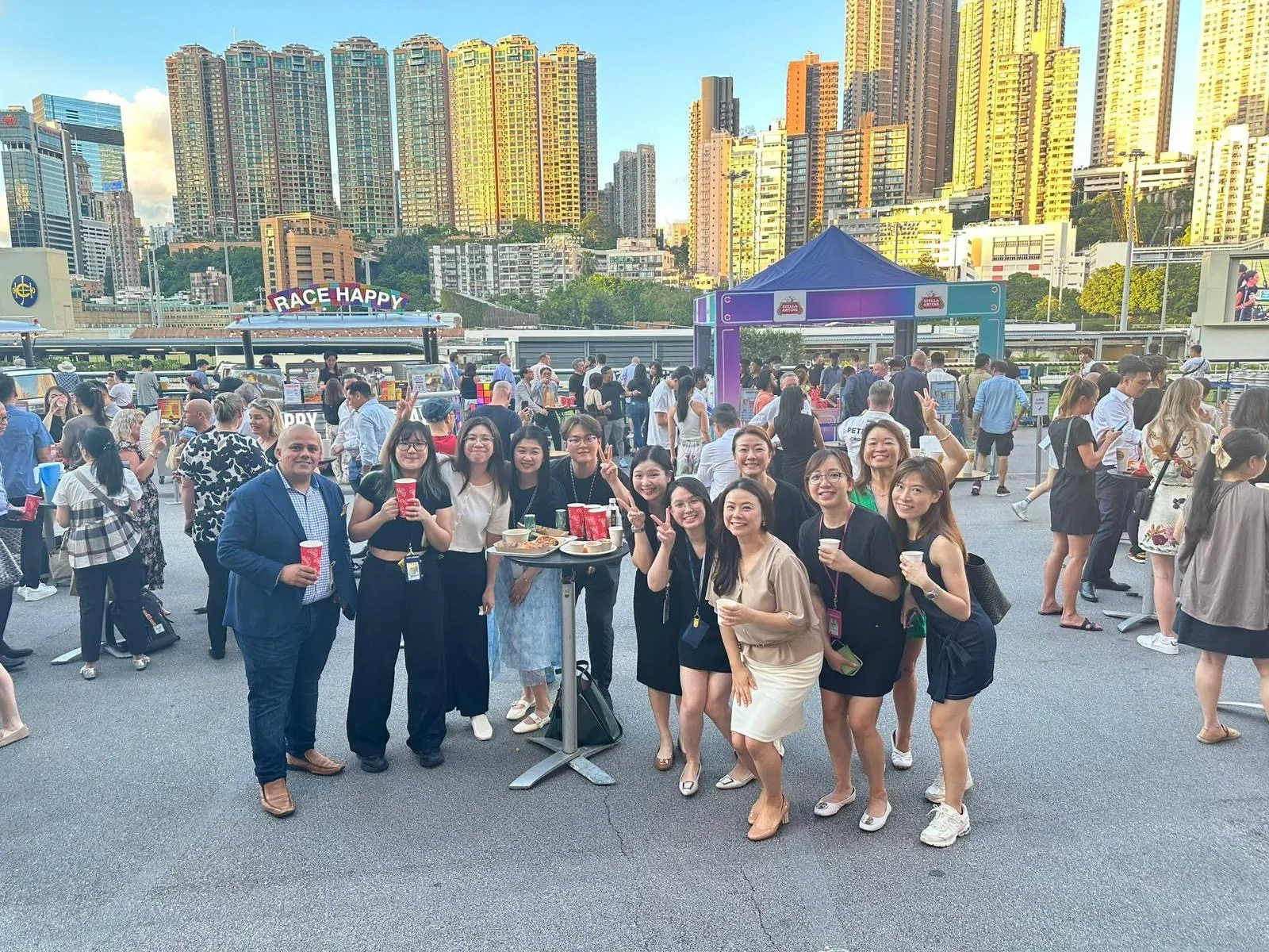 Paul Hotchan and colleagues hold red cups at a bustling outdoor food-truck fair inside the racecourse, Hong Kong’s skyline glowing behind.