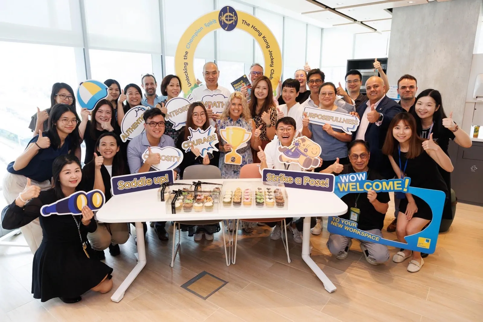 Large mixed team gathers around cupcakes and cartoon speech-bubble props inside a bright office, all flashing thumbs-up beneath a HKJC arch.