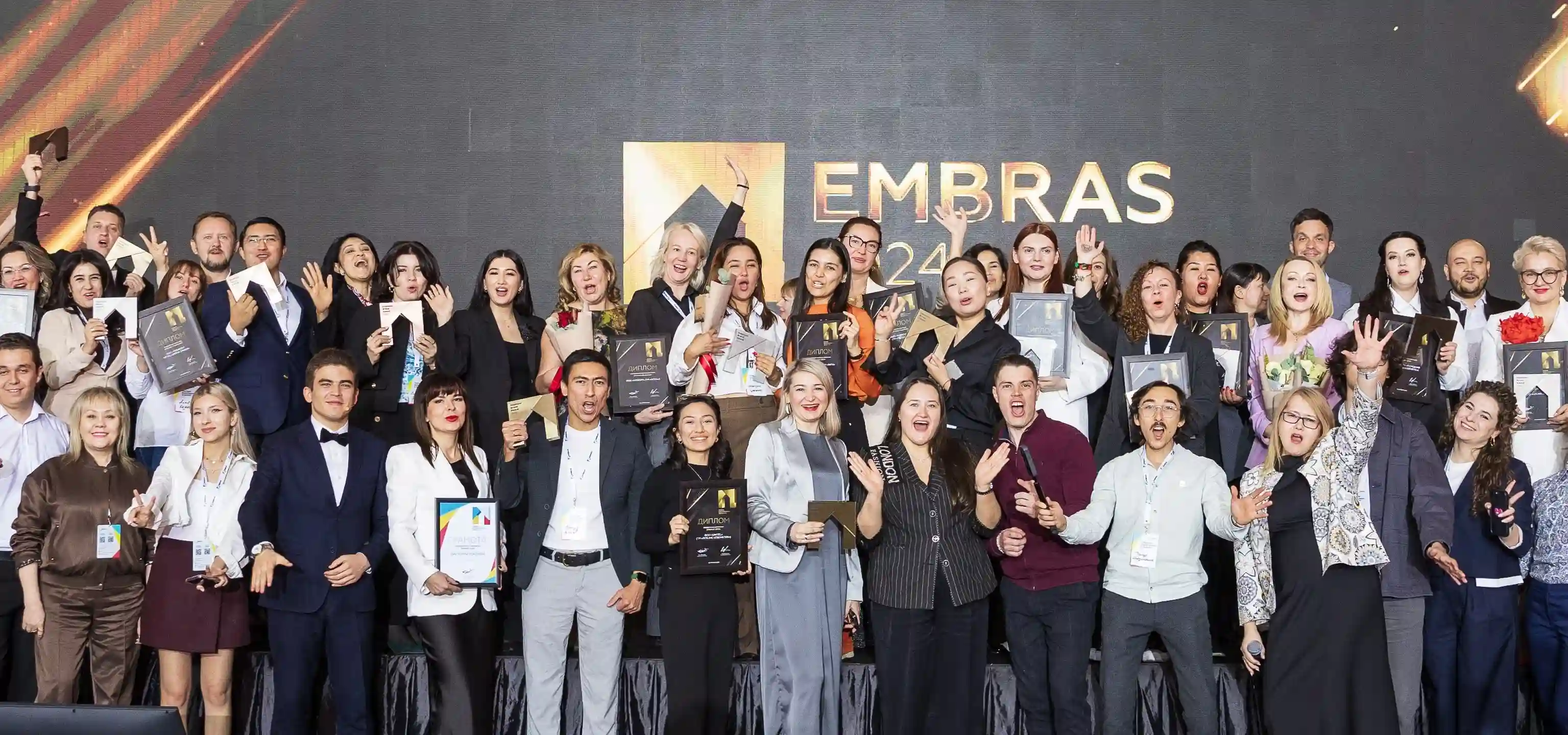 Group of EMBRAS 2024 award winners smiling, cheering, and holding plaques and trophies on stage.