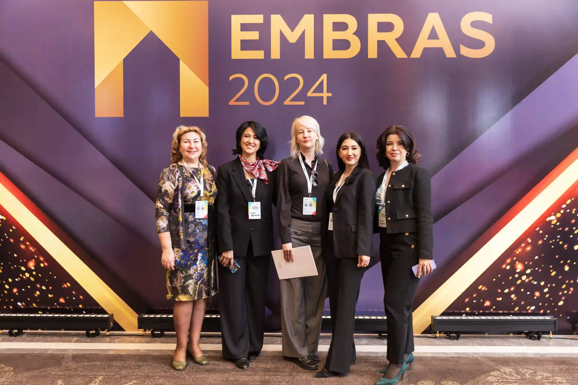 Five formally dressed judges or speakers stand smiling in front of the EMBRAS 2024 backdrop.