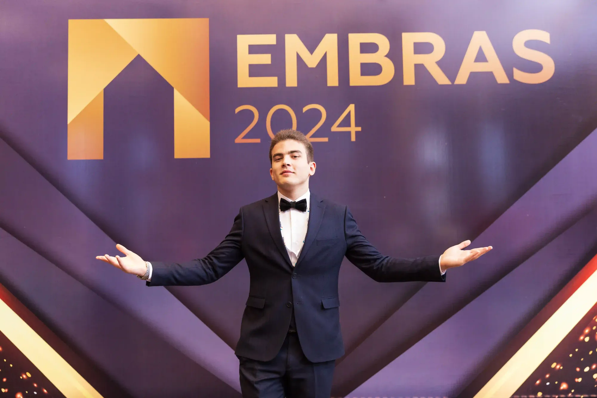 A man in a tuxedo stands with arms wide open in front of the EMBRAS 2024 event backdrop, projecting confidence and celebration.