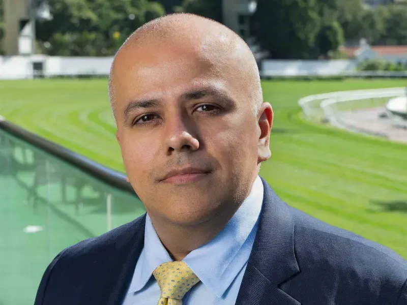 Paul Hotchan in a navy suit and pale-yellow tie stands beside a glass balustrade, lush green racetrack curving in the background under bright daylight.