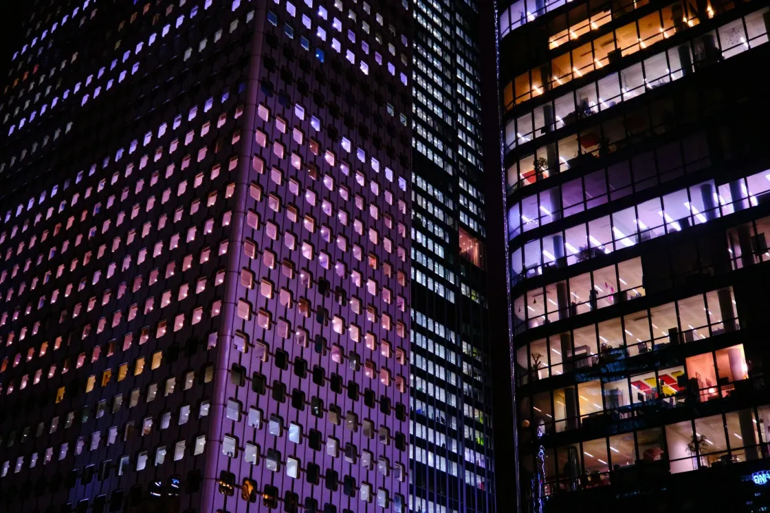 Night view of modern office towers with purple-blue lighting, showing many lit windows and glass-walled offices across several floors.