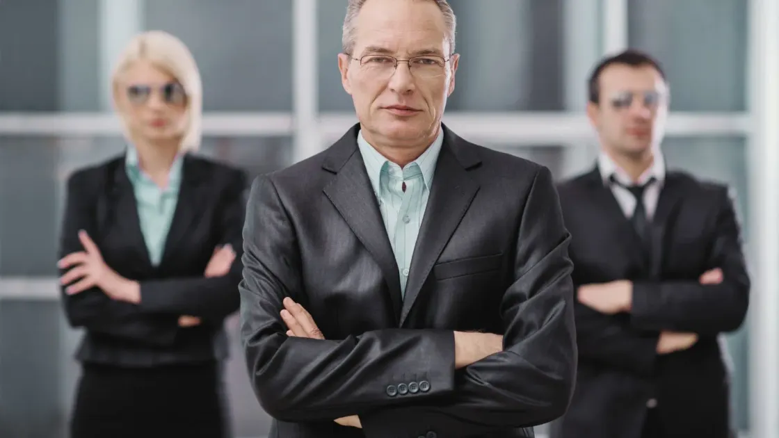Stern executive in a dark suit with arms crossed, two colleagues blurred behind, symbolizing a top-down “agree or quit” workplace ultimatum.