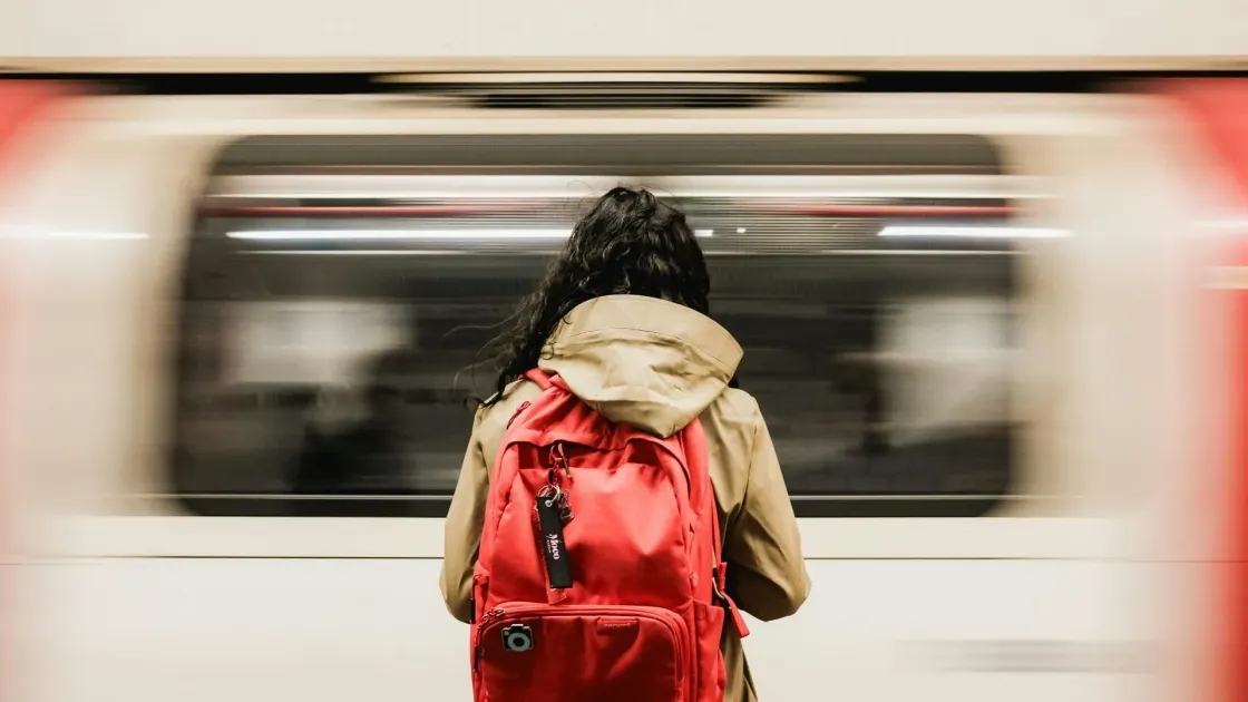 Rear view of a commuter in a tan coat with a bright red backpack standing on a subway platform as a train rushes past in motion blur.
