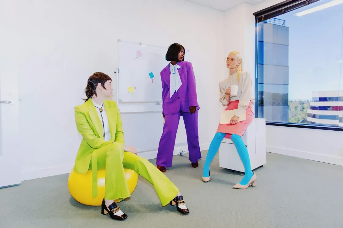 Three colleagues in a bright office space wearing bold, fashion-forward workwear, standing and seated near a whiteboard and large window.