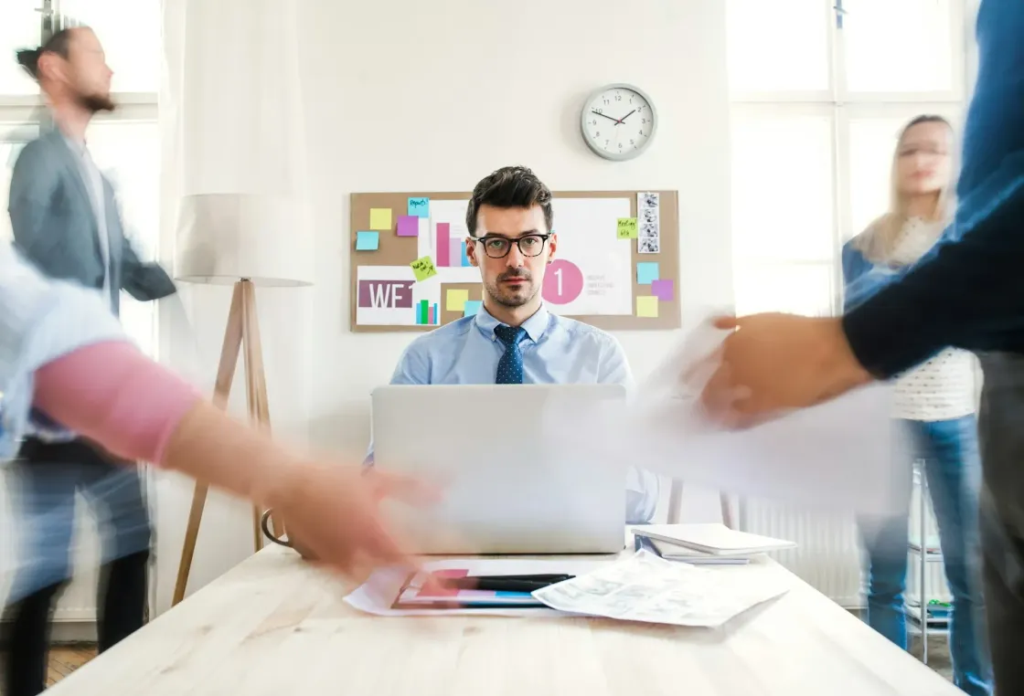 Office worker sitting at a laptop while blurred colleagues and papers move around him, suggesting workload pressure and work intensification.