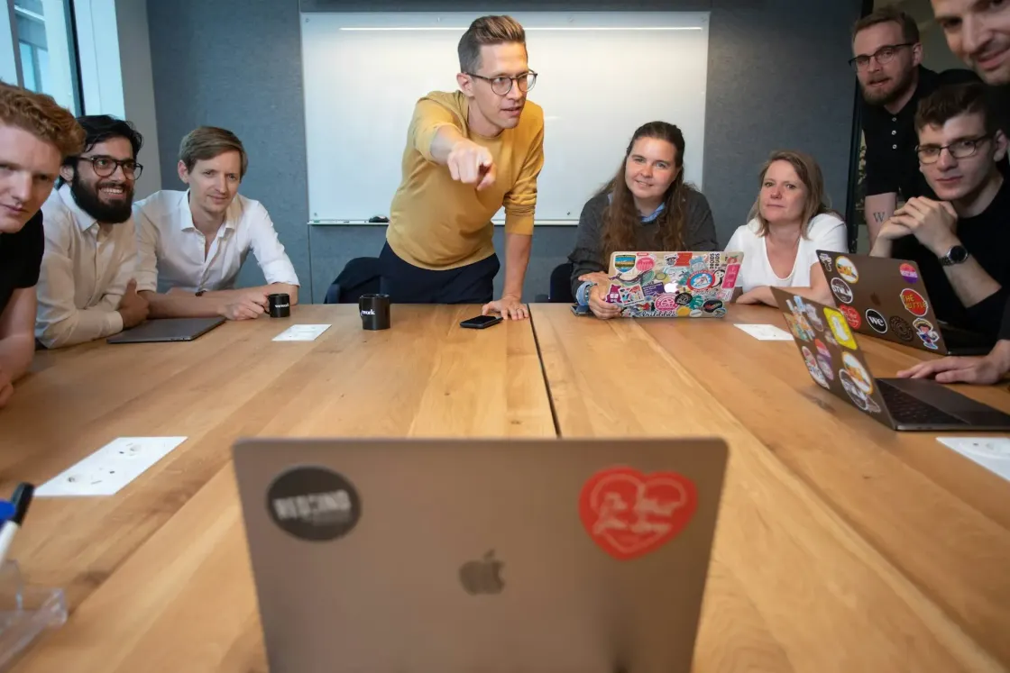 A team gathered around a meeting room table, one person leaning forward and pointing, laptops open; the moment just before someone asks what the brief actually says.