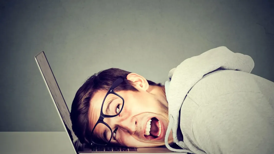 A person in glasses lying face-down on a laptop keyboard with their mouth open in frustration, suggesting burnout and work overload.
