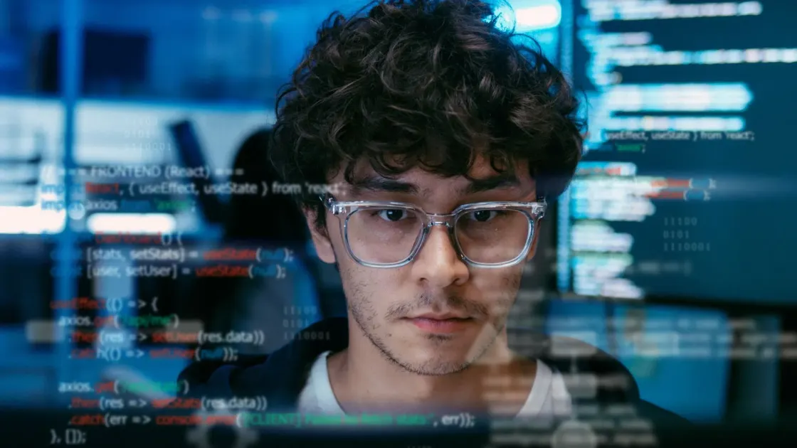 Close-up of a young professional wearing glasses with lines of computer code reflected across the image, suggesting AI-driven work, pressure, and scrutiny.