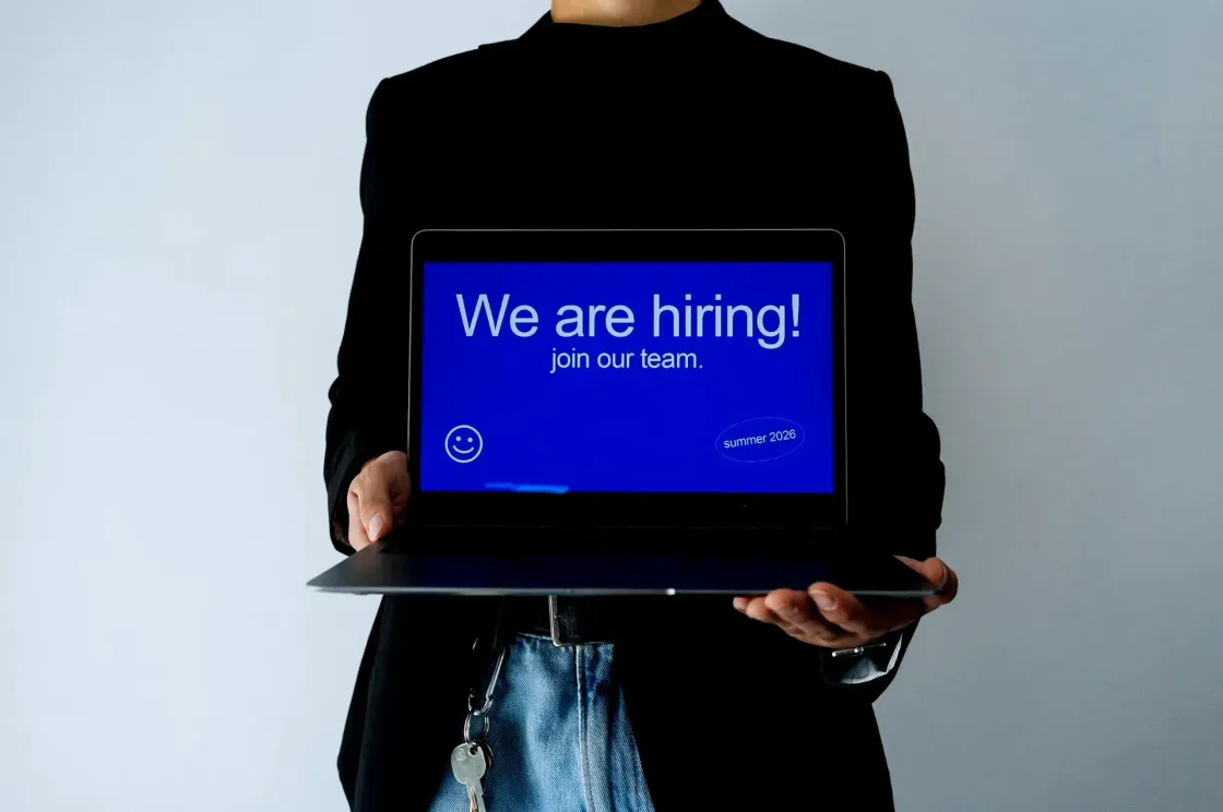 A person holds an open laptop displaying a bold blue "We are hiring, join our team" screen, representing recruitment marketing as the visible, campaign-driven layer of talent attraction.