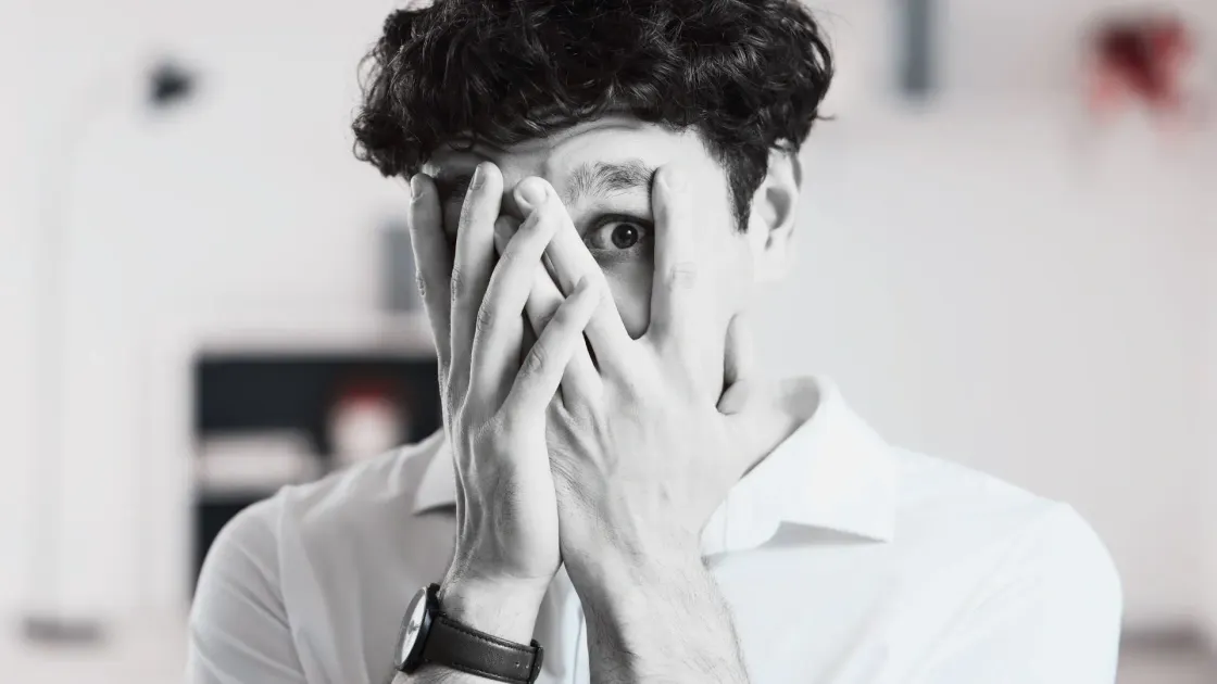 A young person covering most of their face with their hands, one eye visible, conveying anxiety, overwhelm, or apprehension in response to workplace change.